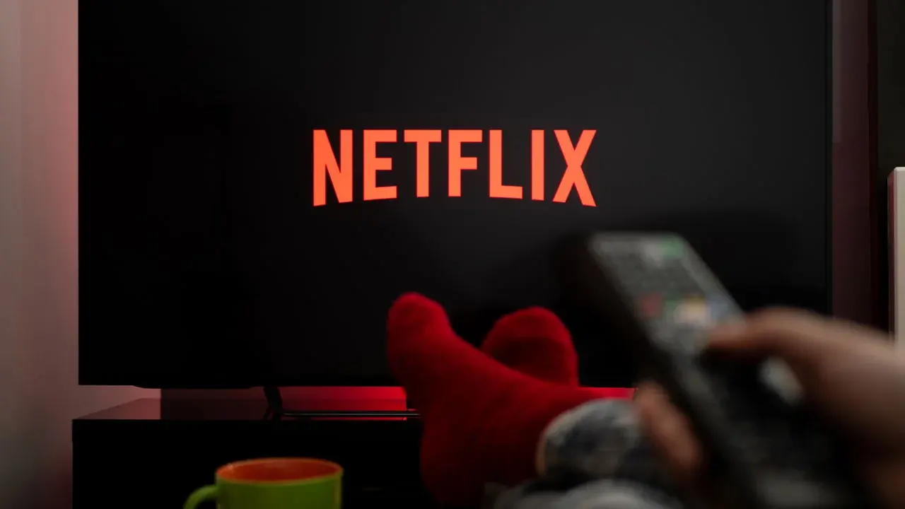 Tela de televisão exibindo o logotipo da Netflix, com uma pessoa segurando um controle remoto em primeiro plano.
